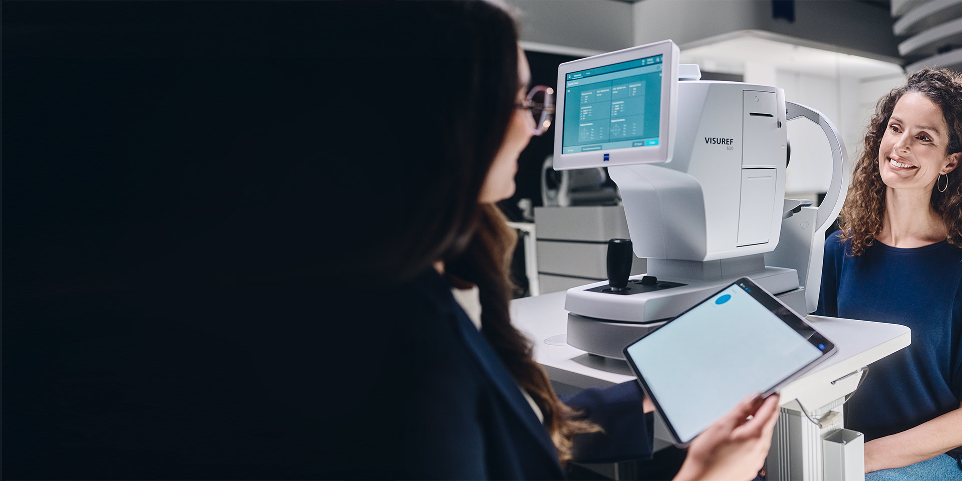 Une opticienne assise devant le ZEISS VISUREF 600 regarde sa tablette et discute avec son client.