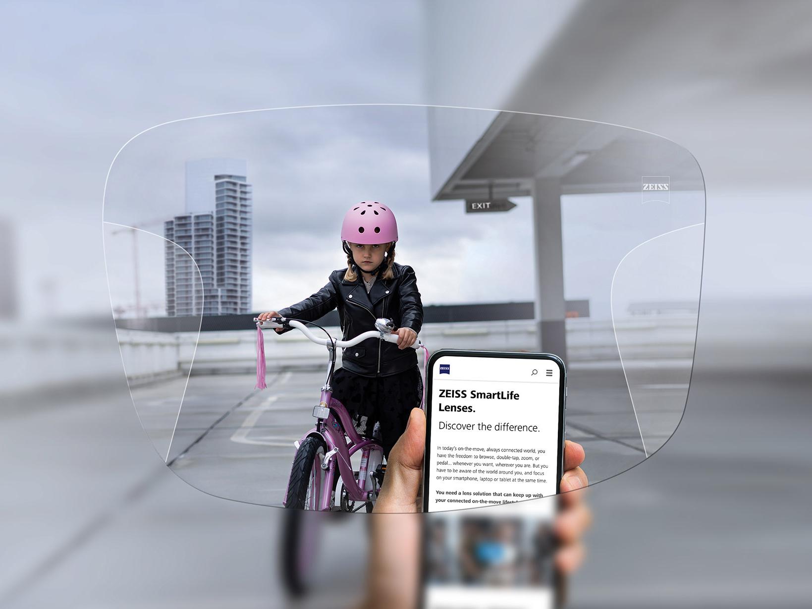 Une personne regarde son téléphone à travers des verres progressifs ZEISS SmartLife. Une petite fille sur son vélo avec un casque rose est visible en arrière-plan. 