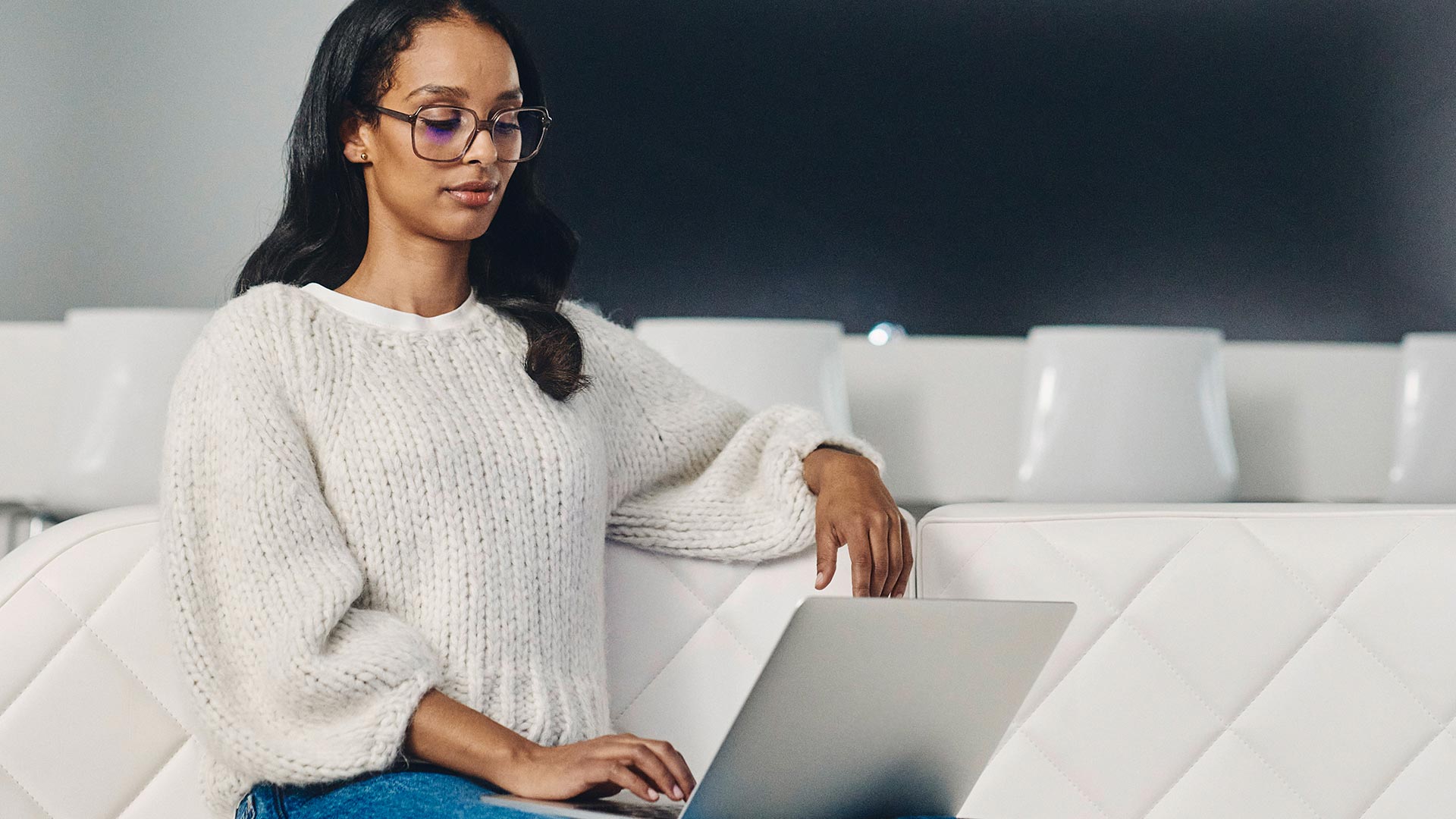 Donkerharige vrouw met donkere bril en een witte trui kijkt op een laptop.