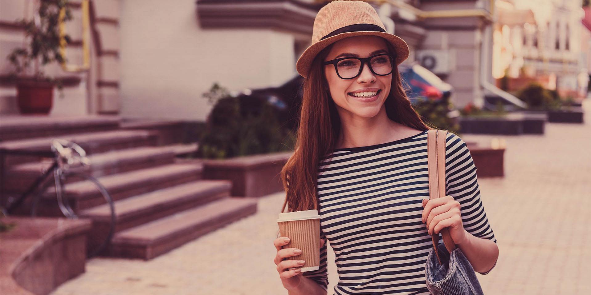 Une jeune femme avec une casquette et des lunettes porte une tasse à café (à droite) et un sac sur l&apos;épaule gauche.