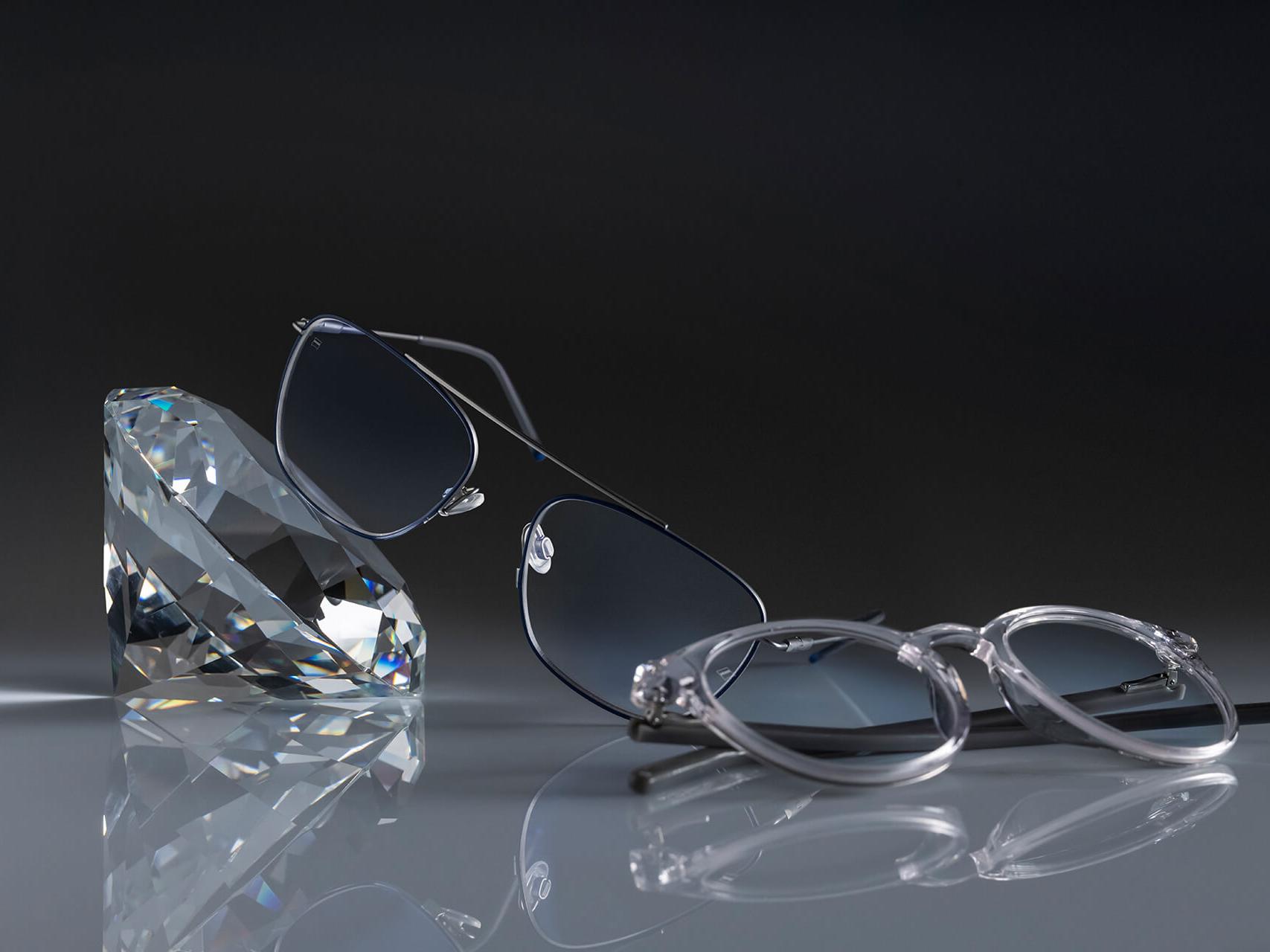Twee brillen met ZEISS brillenglazen met de hardste ZEISS coating ooit - DuraVision® Platinum. Eén bril leunt tegen een kristal, de andere ligt plat op de grond. Beide hebben heldere brillenglazen zonder blauwachtige reflectie.