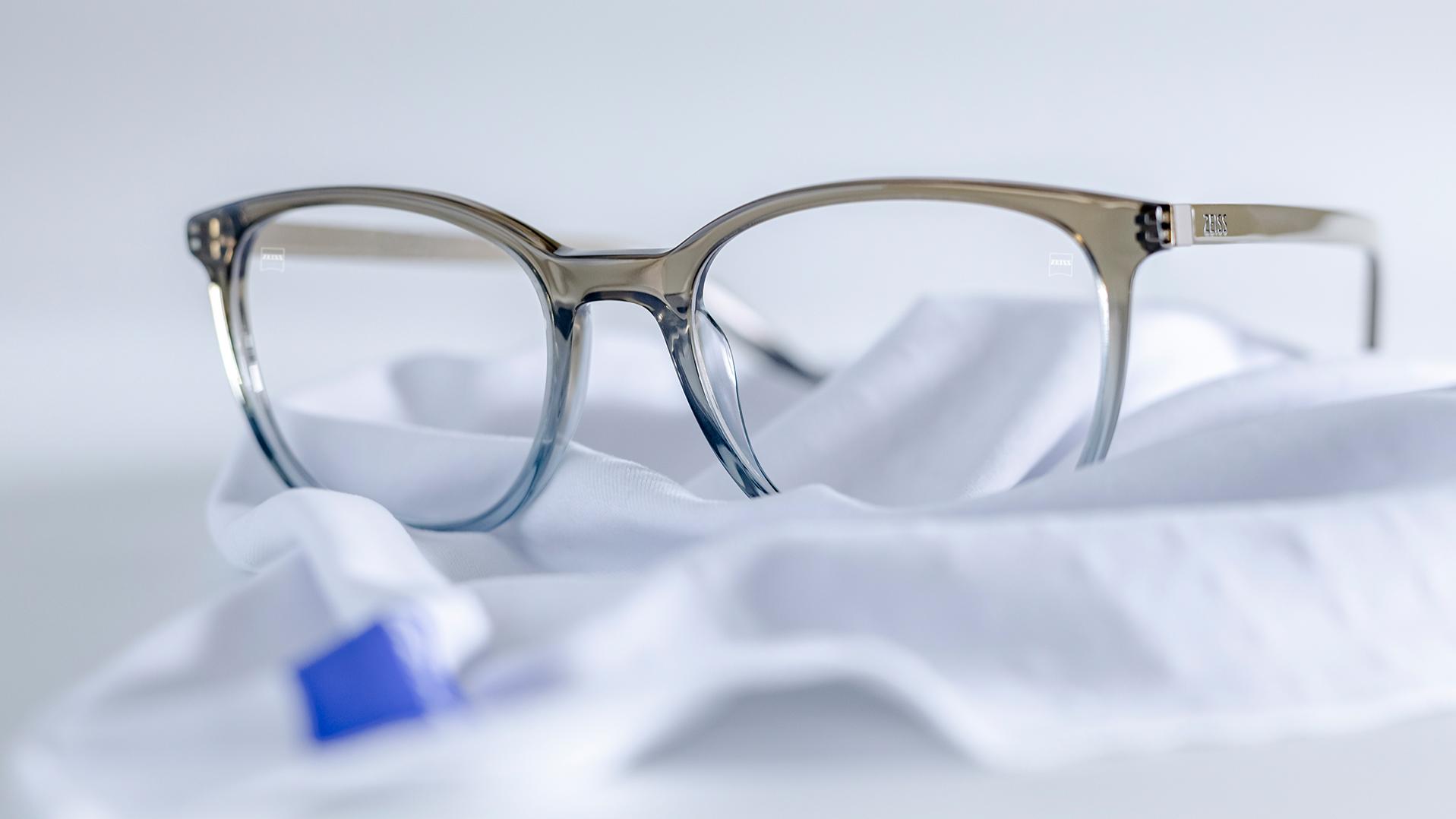 Een bril met grijsblauw montuur en ZEISS brillenglazen met DuraVision® coating ligt op een wit microvezeldoekje.