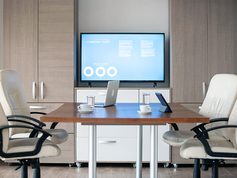 Uitzicht vanaf het bureau van een directiekantoor. Een paar meter verderop is een monitor op een muur gemonteerd. Er staat een kleine vergadertafel met bureaustoelen en laptops op tafel.