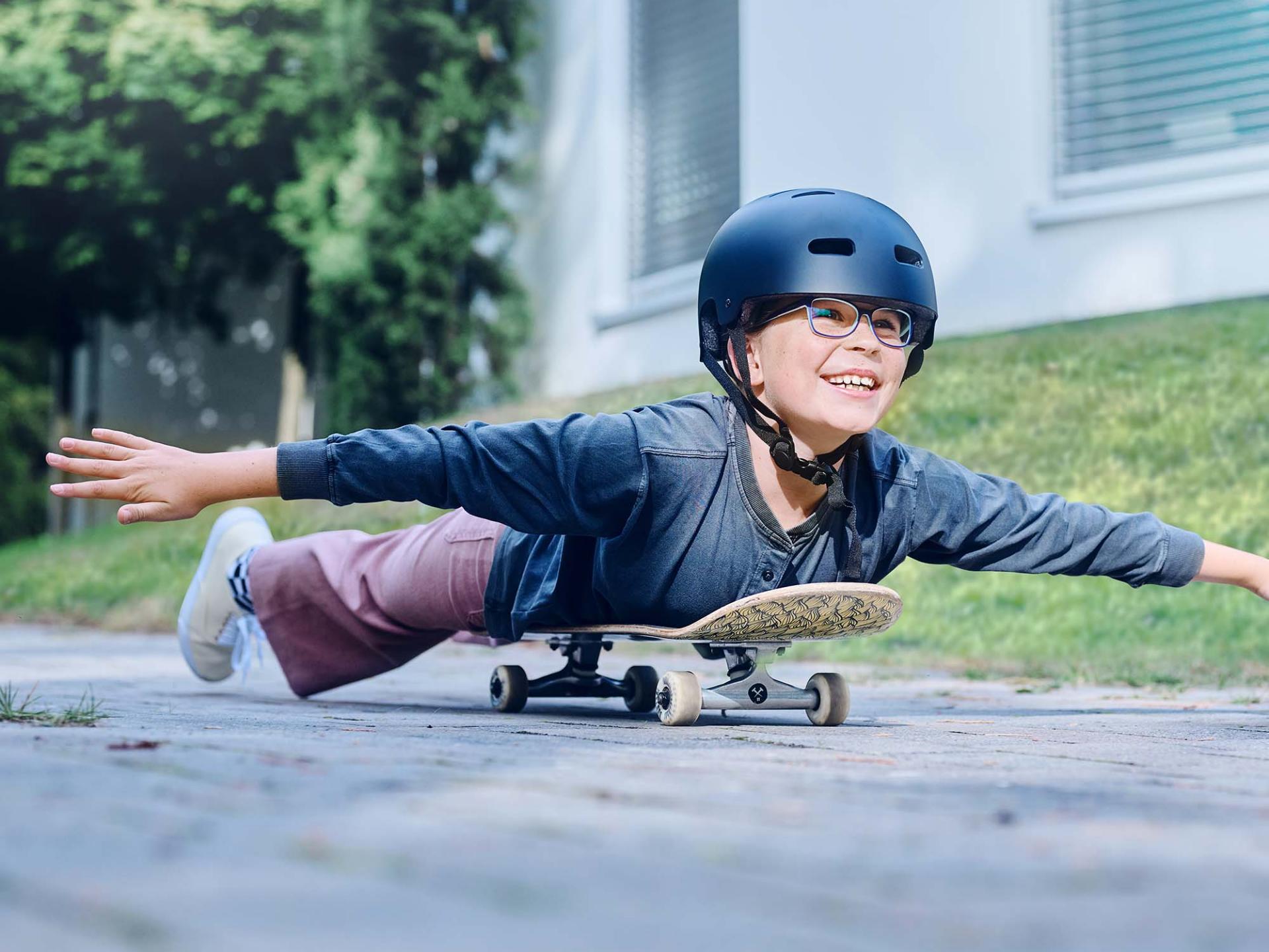 Een meisje met een helm en een bril rolt liggend op een skateboard de weg af en strekt haar armen uit.